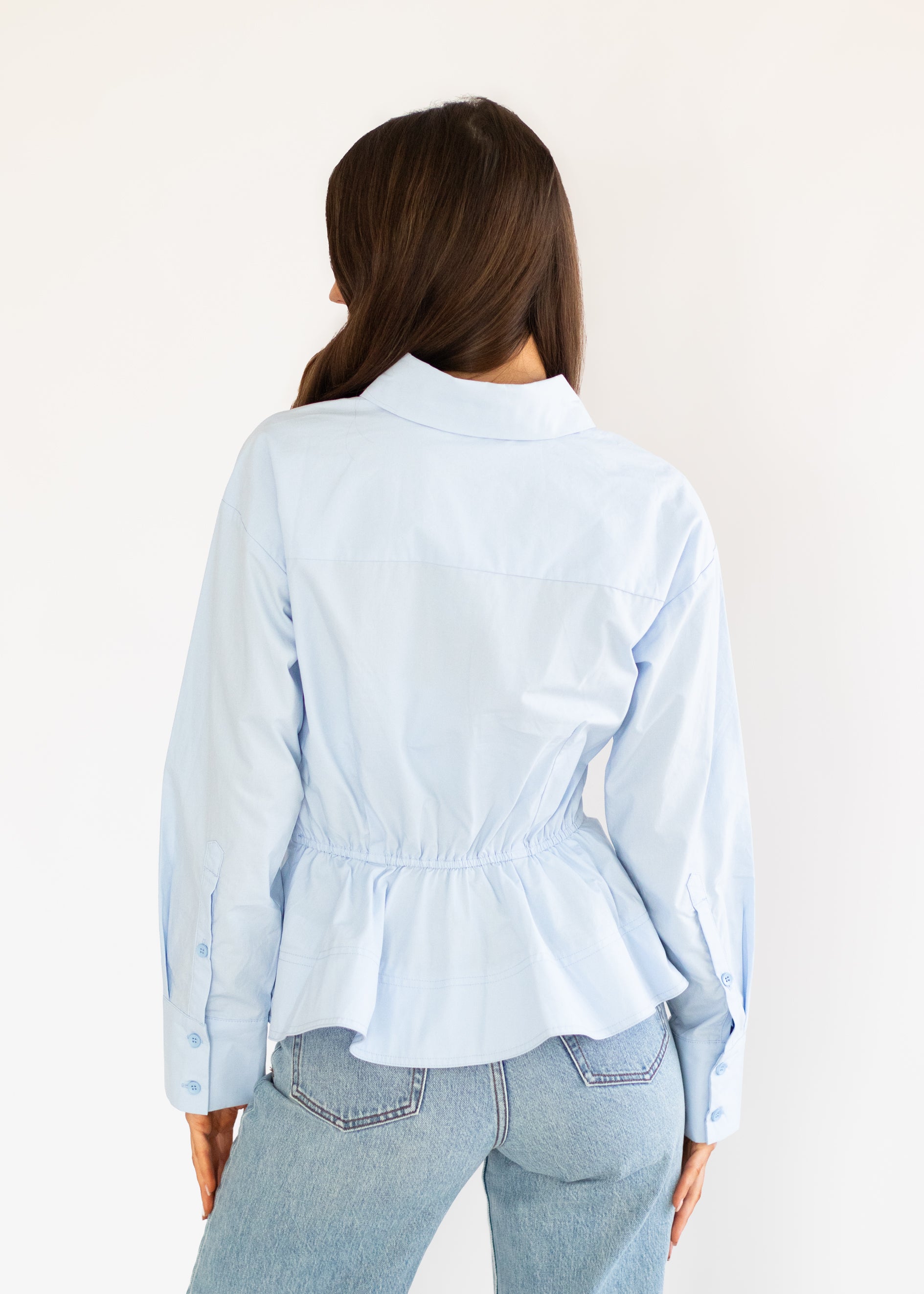 A woman viewed from behind wearing a light blue long-sleeve button-up shirt with a gathered peplum waist and light-wash blue jeans, set against a plain off-white background.
