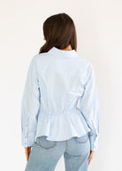 A woman viewed from behind wearing a light blue long-sleeve button-up shirt with a gathered peplum waist and light-wash blue jeans, set against a plain off-white background.