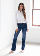 A woman modeling dark blue straight-leg jeans with a white button-down shirt, standing in a bright, white room.