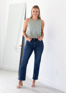 A woman modeling a sleeveless sage green top with a subtle floral pattern and dark blue cropped jeans, standing in a bright, minimalist room with a large mirror on the left.