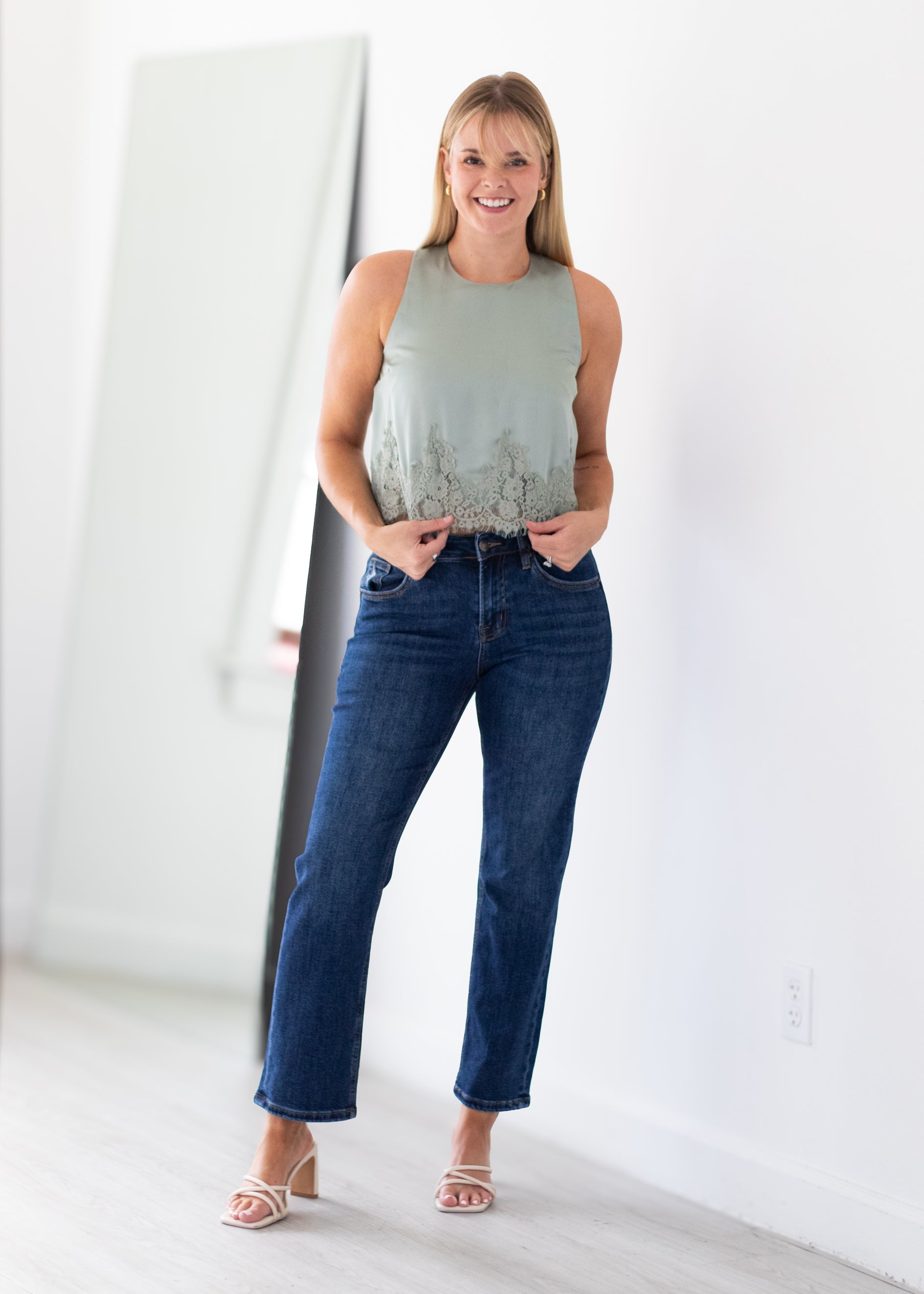 A woman modeling a sleeveless sage green top with a subtle floral pattern and dark blue cropped jeans, standing in a bright, minimalist room with a large mirror on the left.