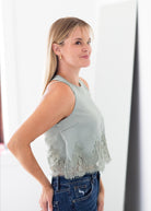 A woman wearing a sleeveless sage green top with a lace hem detail, paired with blue jeans, standing in a bright indoor setting.