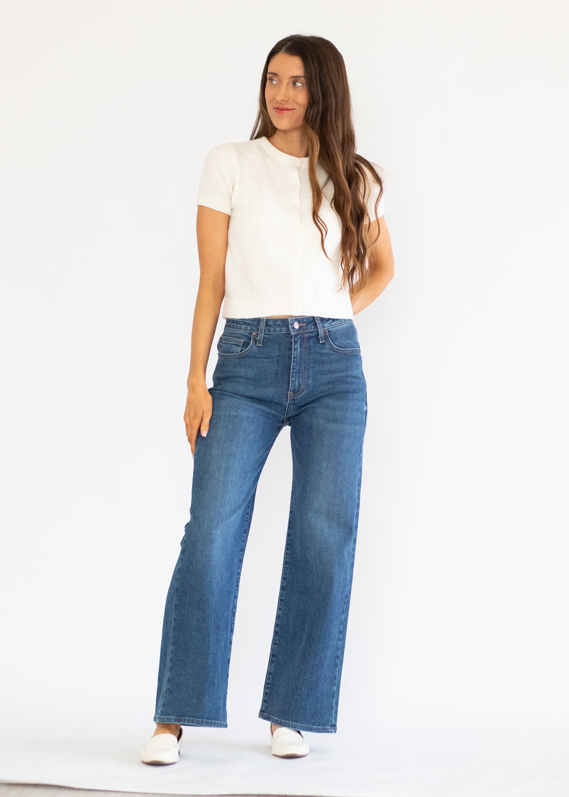 A woman modeling a pair of high-waisted, wide-leg blue jeans with a white short-sleeve t-shirt and white shoes, standing against a plain white studio background.