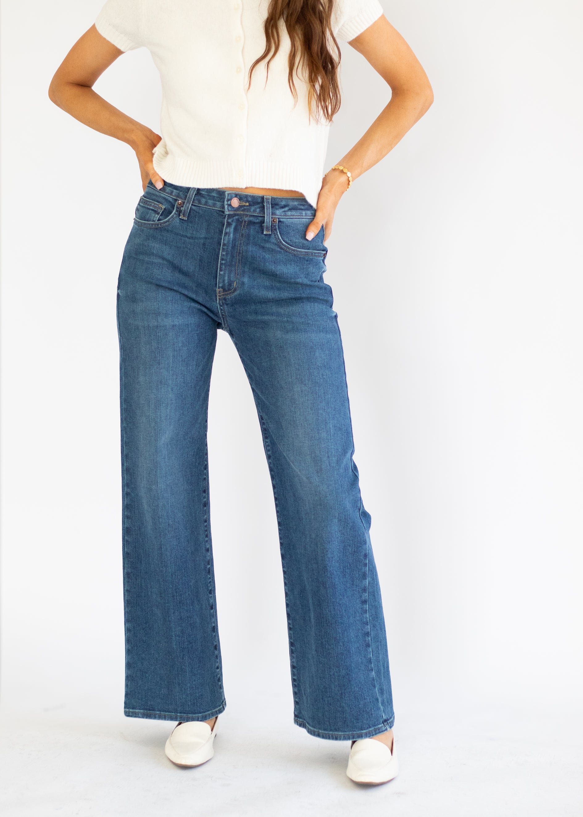 A model wearing a pair of high-waisted, wide-leg blue denim jeans with a straight-to-flared leg, paired with a white top and white shoes, standing against a plain white studio background.
