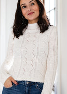 Woman wearing a white cable knit sweater with a blurred indoor background