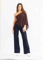 A woman modeling a dark brown one-shoulder draped top with dark blue wide-leg jeans and white shoes, standing against a plain white studio background.