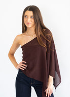 A woman modeling a dark brown, one-shoulder draped wrap top or poncho over dark blue jeans, set against a plain white studio background.