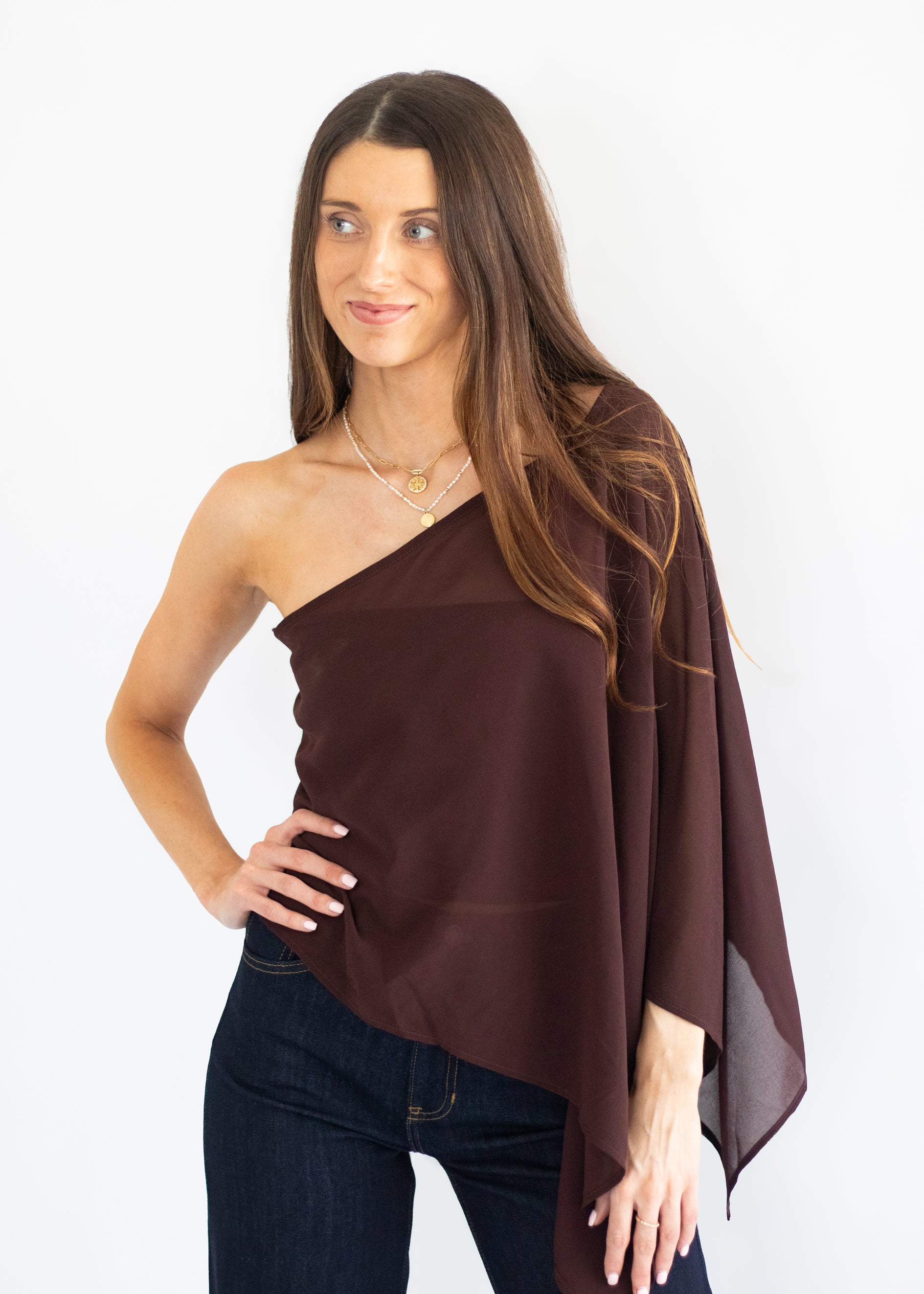 A woman modeling a dark brown, one-shoulder draped wrap top or poncho over dark blue jeans, set against a plain white studio background.