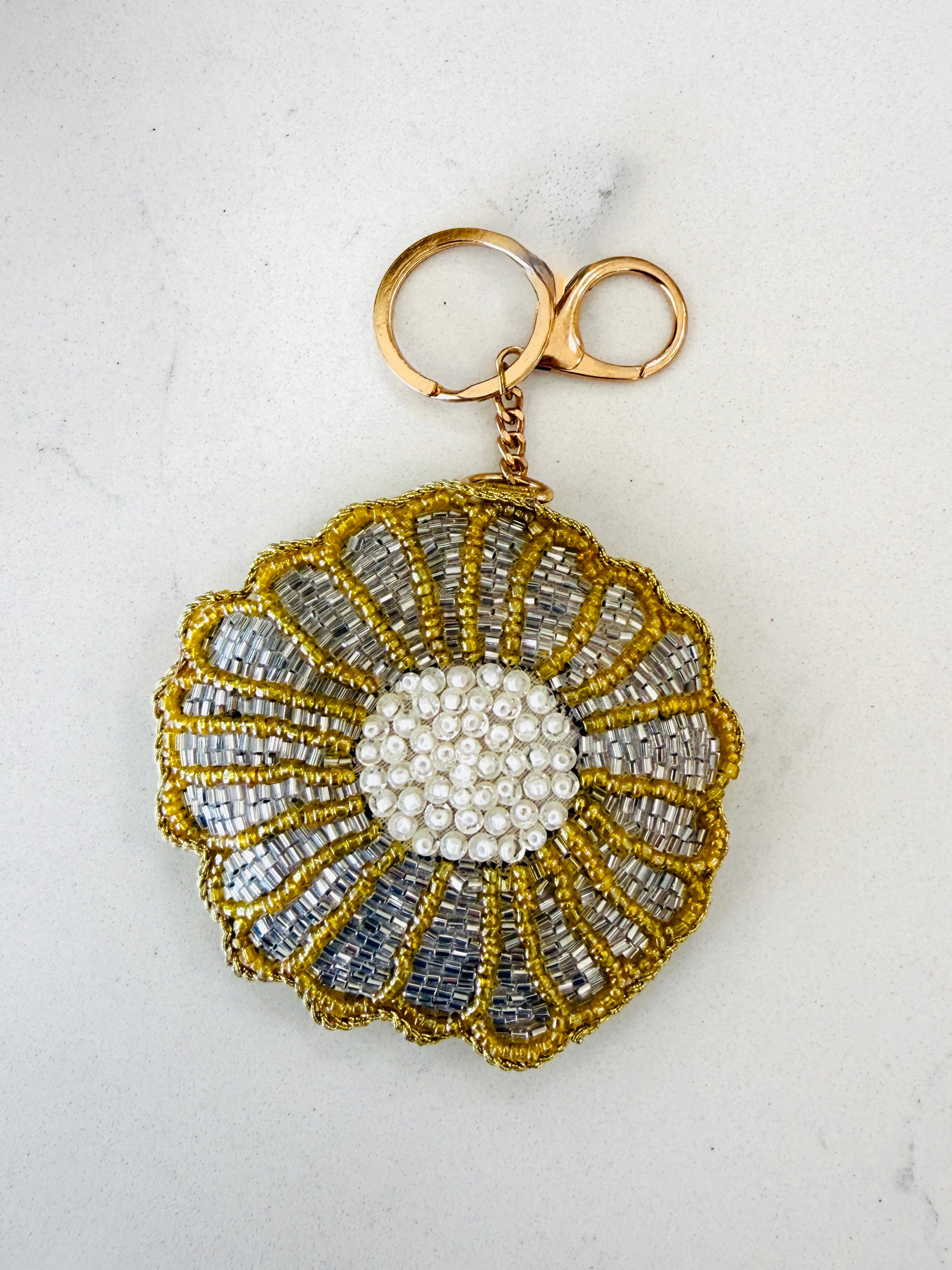 A handmade beaded flower-shaped keychain with a gold-tone clasp, featuring a white beaded center and gold and silver beaded petals, set against a plain off-white background.