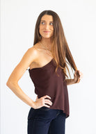 A woman modeling a dark brown, one-shoulder satin-like top paired with dark blue jeans, set against a plain white studio background.