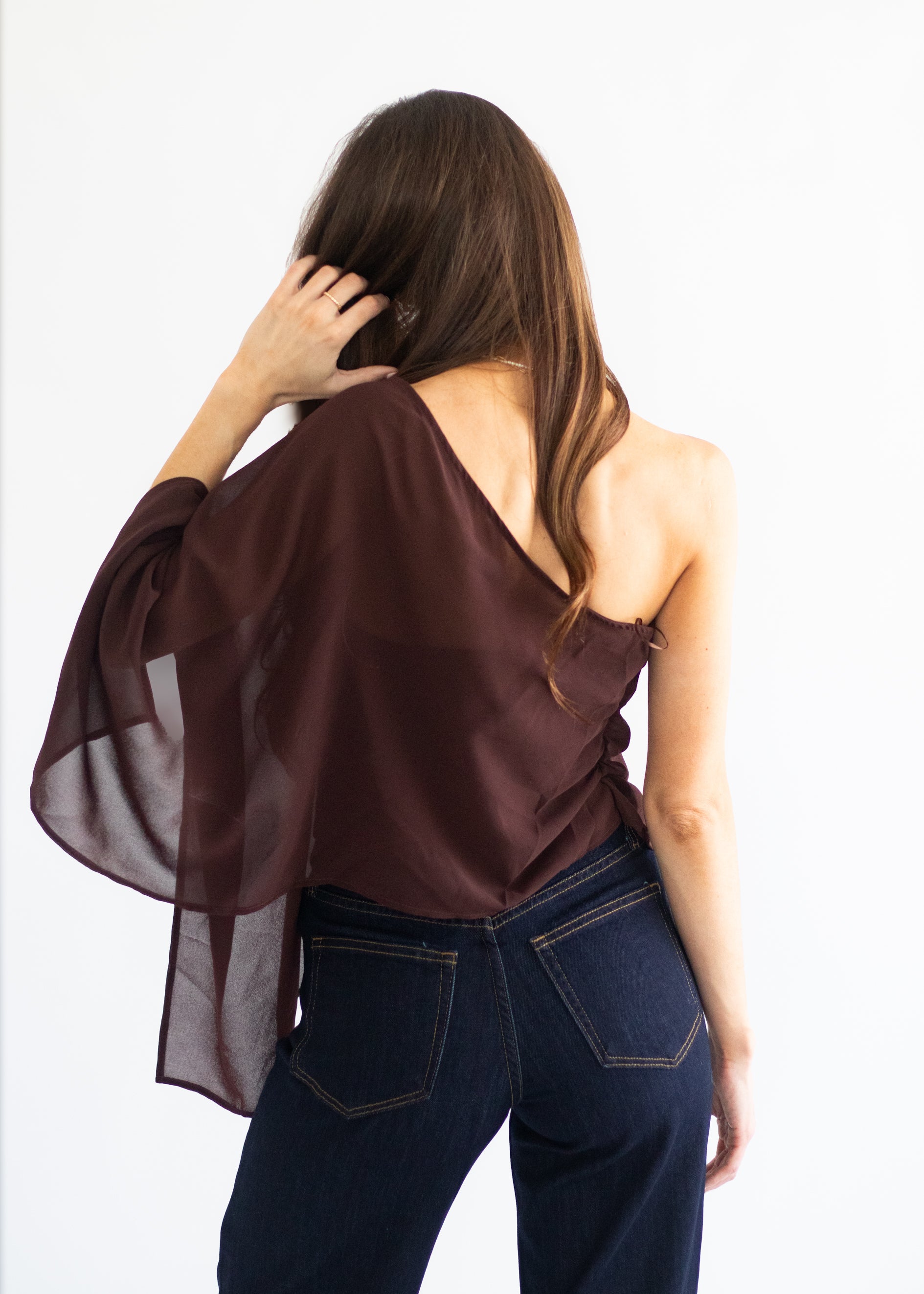 A woman shown from the back wearing a sheer, one-shoulder draped top in a deep plum-brown color, paired with dark blue jeans, against a plain white background.