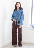 A woman modeling a light blue denim belted jacket and dark brown wide-leg pants in a bright, white-walled room.