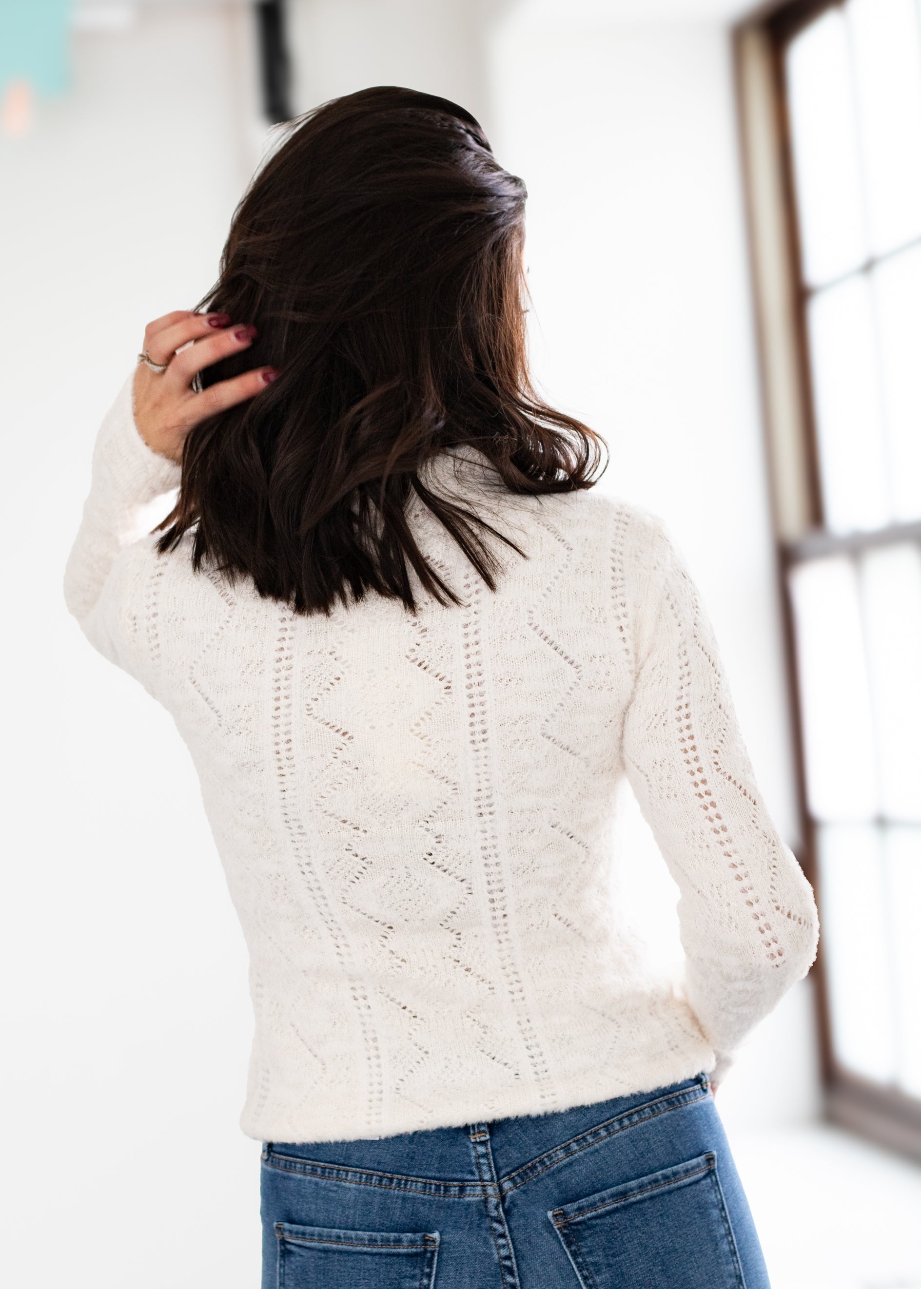 Person wearing a white cable knit sweater and blue jeans, standing indoors.