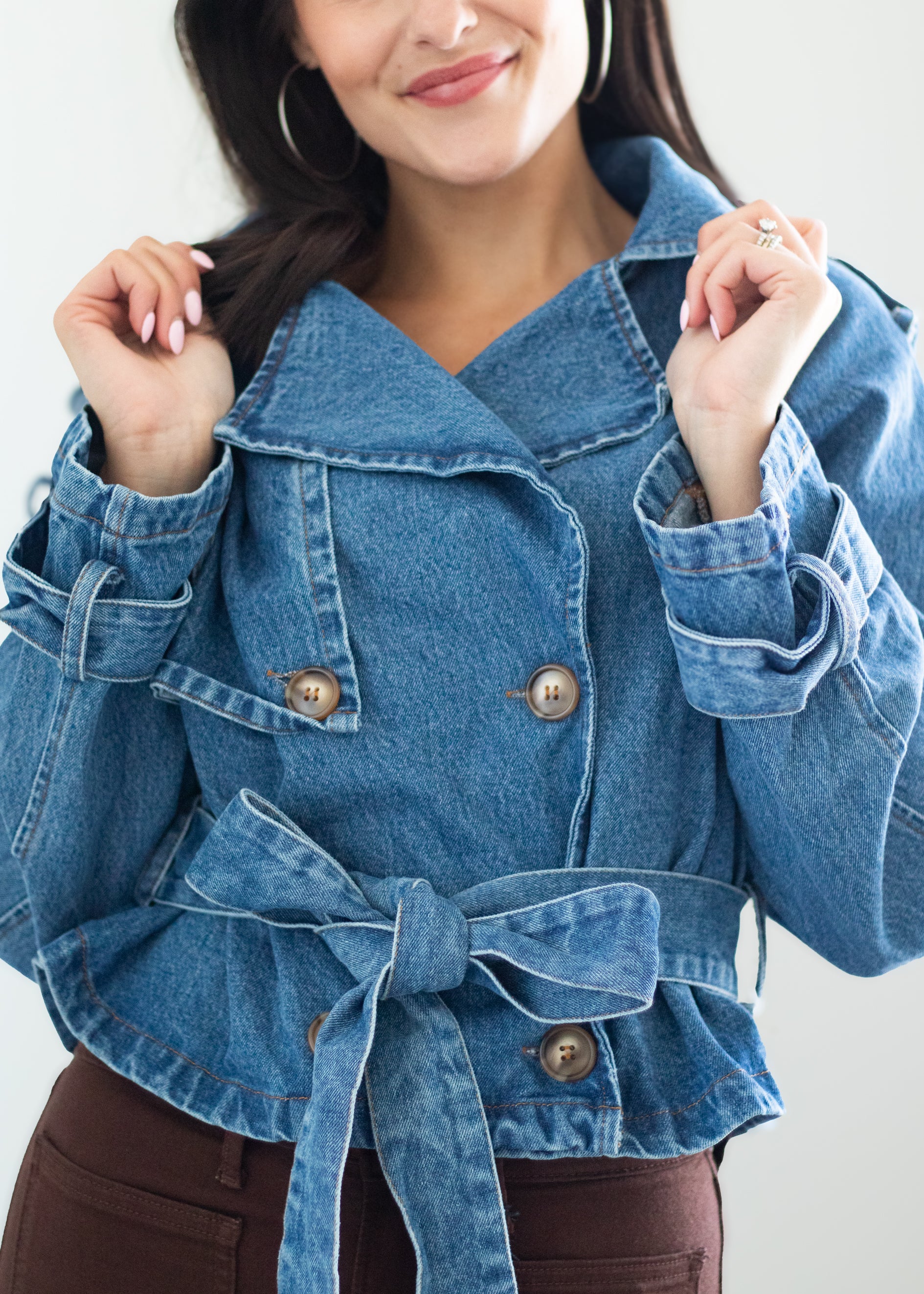 A woman wearing a cropped blue denim jacket with a belted waist and double-breasted buttons, paired with brown pants, against a plain light background.