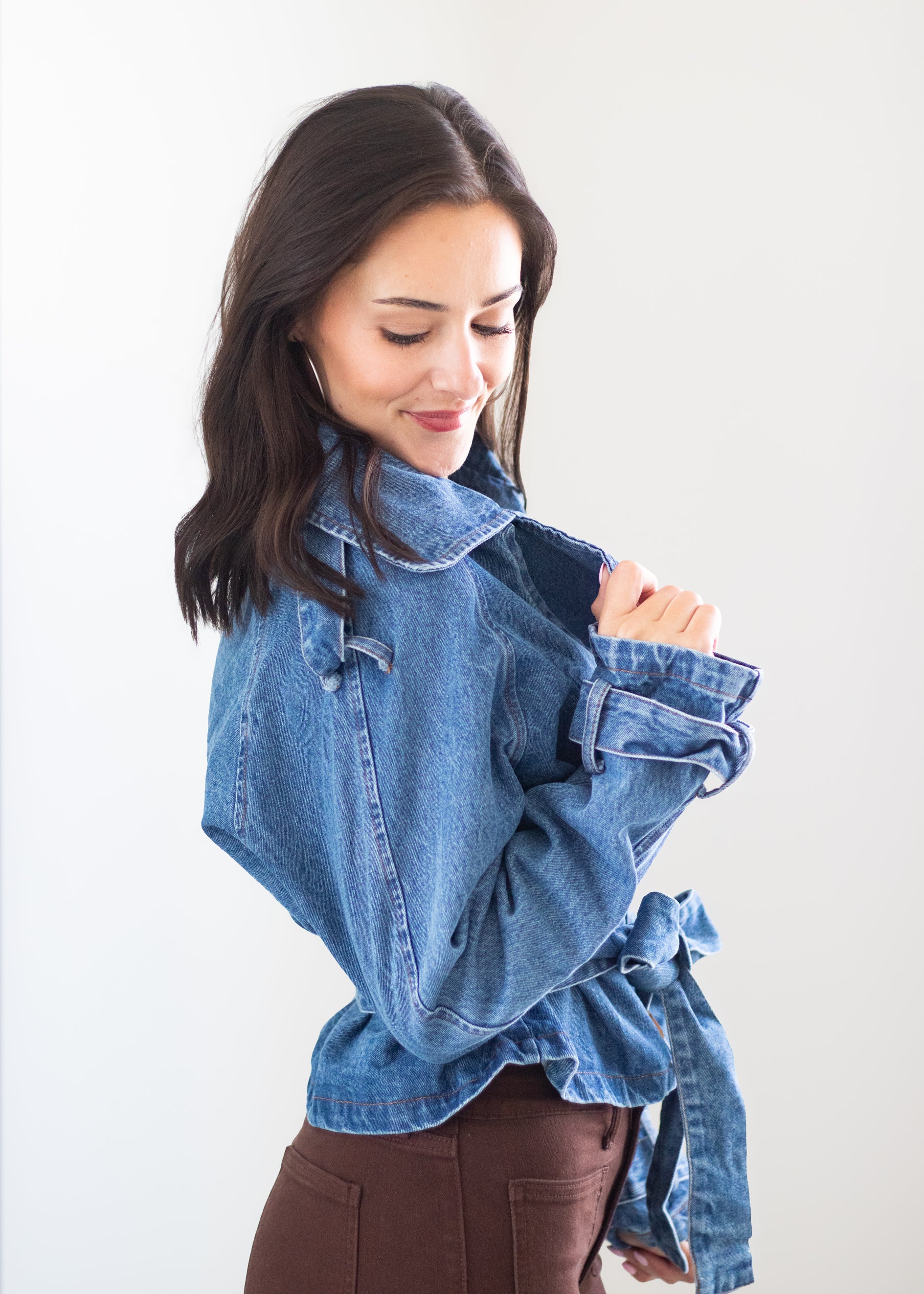 A woman wearing a light blue denim jacket with a tie waist and rolled sleeves, paired with brown pants, posing against a plain white background.