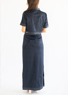 A woman viewed from the back wearing a dark navy blue short-sleeve collared shirt dress with a matching belt and long skirt, set against a plain white studio background.