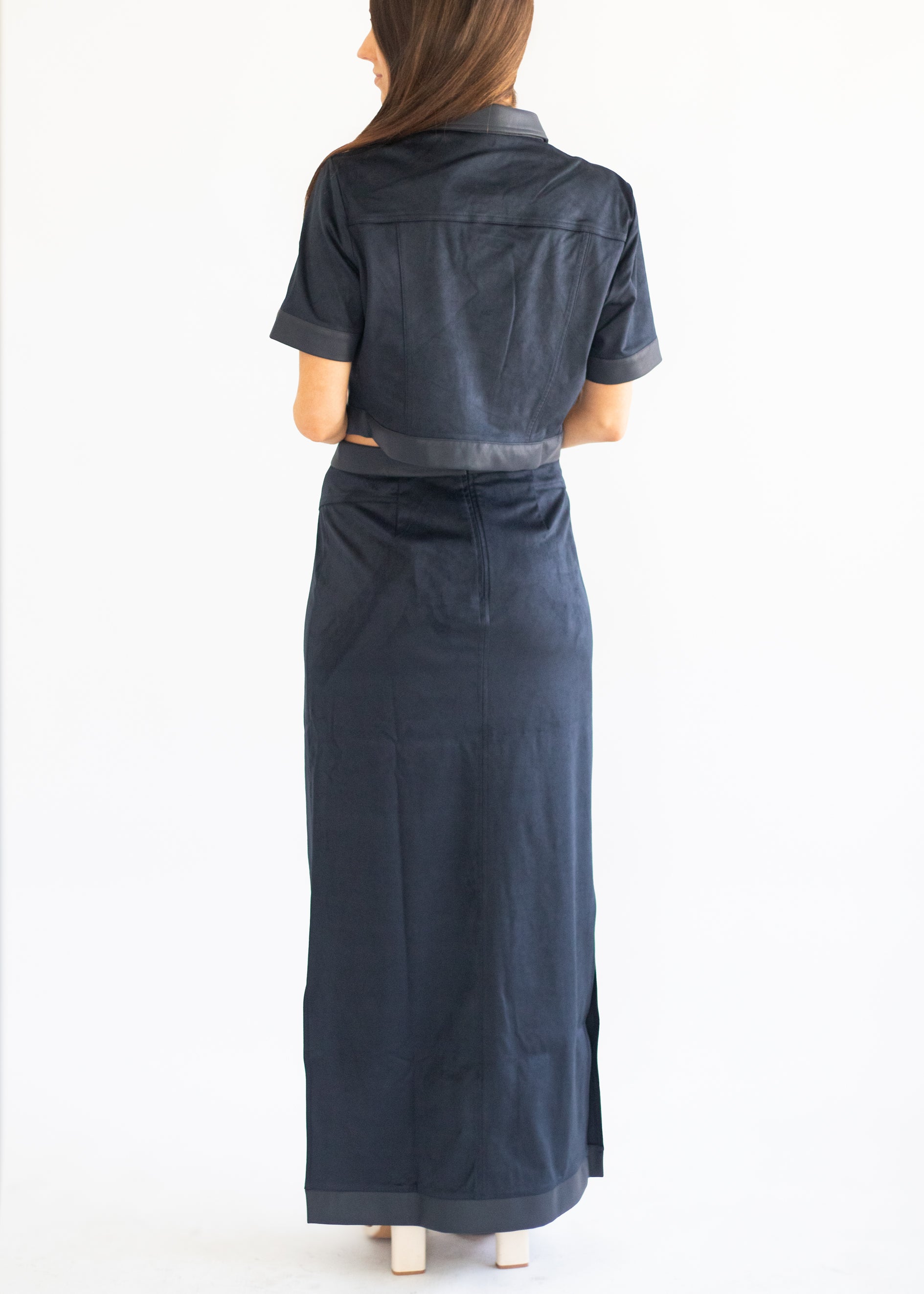A woman viewed from the back wearing a dark navy blue short-sleeve collared shirt dress with a matching belt and long skirt, set against a plain white studio background.