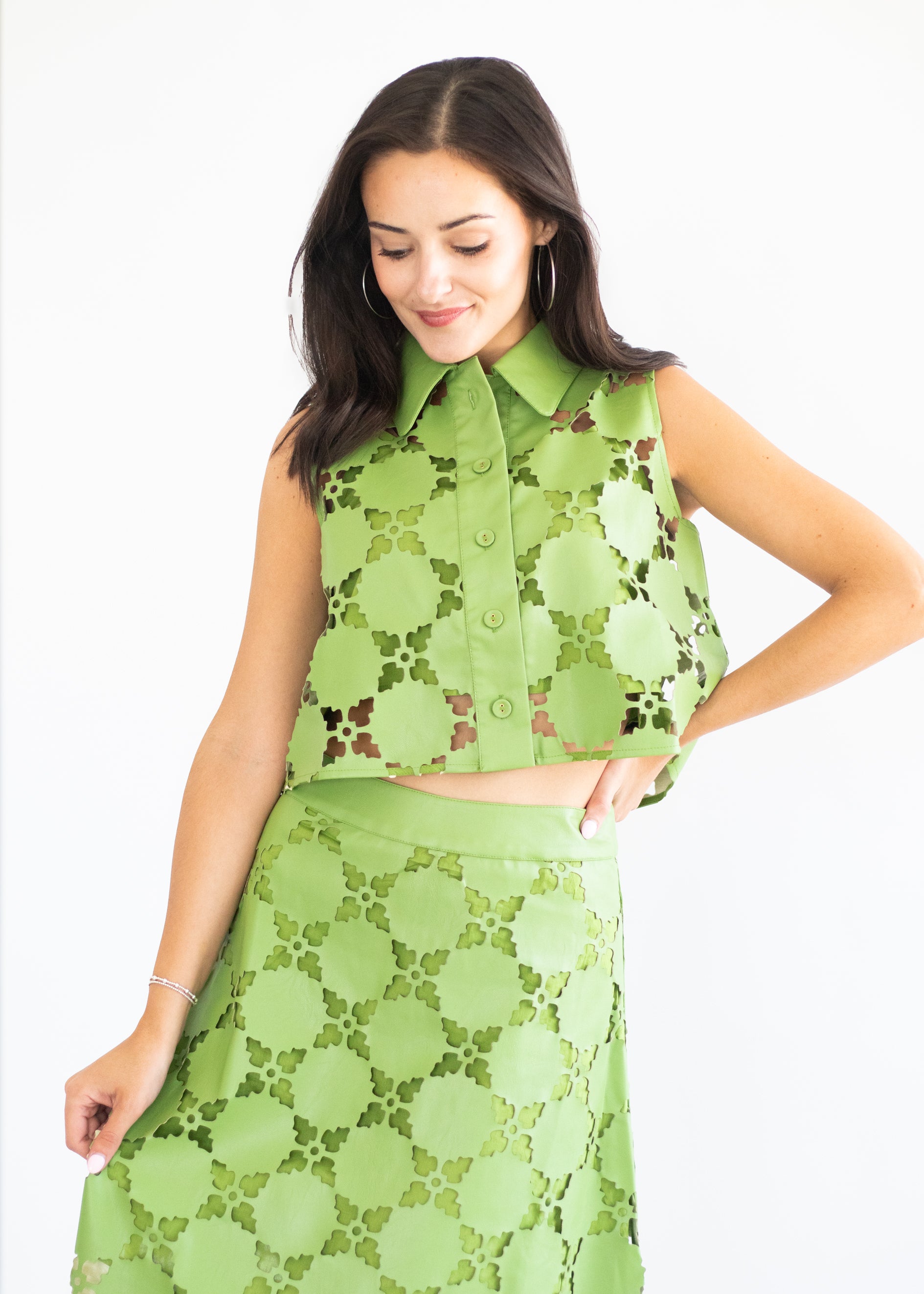 A woman modeling a matching two-piece green lace set consisting of a sleeveless collared button-front top and a high-waisted skirt, set against a plain white background.