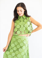A woman modeling a matching two-piece green lace set consisting of a sleeveless collared button-front top and a high-waisted skirt, set against a plain white background.