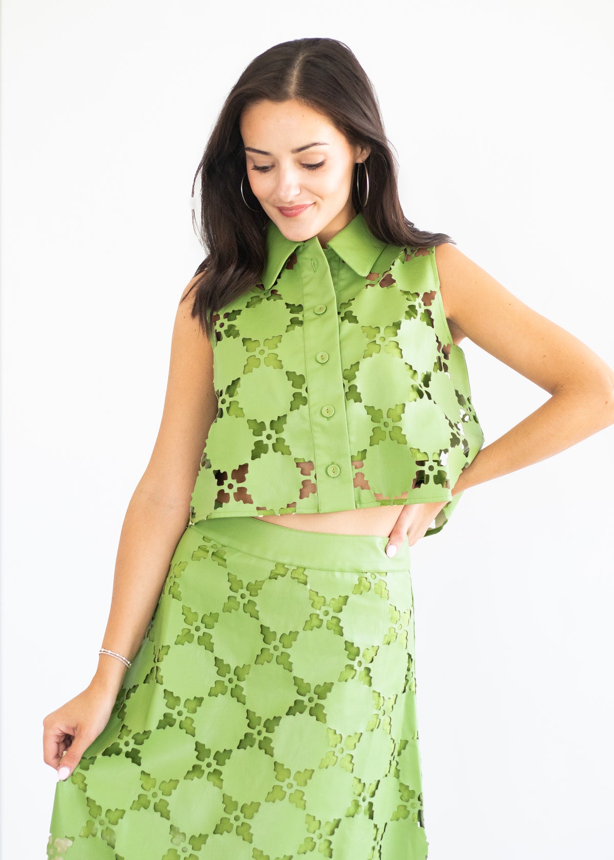 A woman modeling a matching two-piece green lace set consisting of a sleeveless collared button-front top and a high-waisted skirt, set against a plain white background.