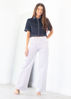 A woman modeling a dark navy short-sleeve button-up shirt and white wide-leg pants, standing in a bright white studio setting.
