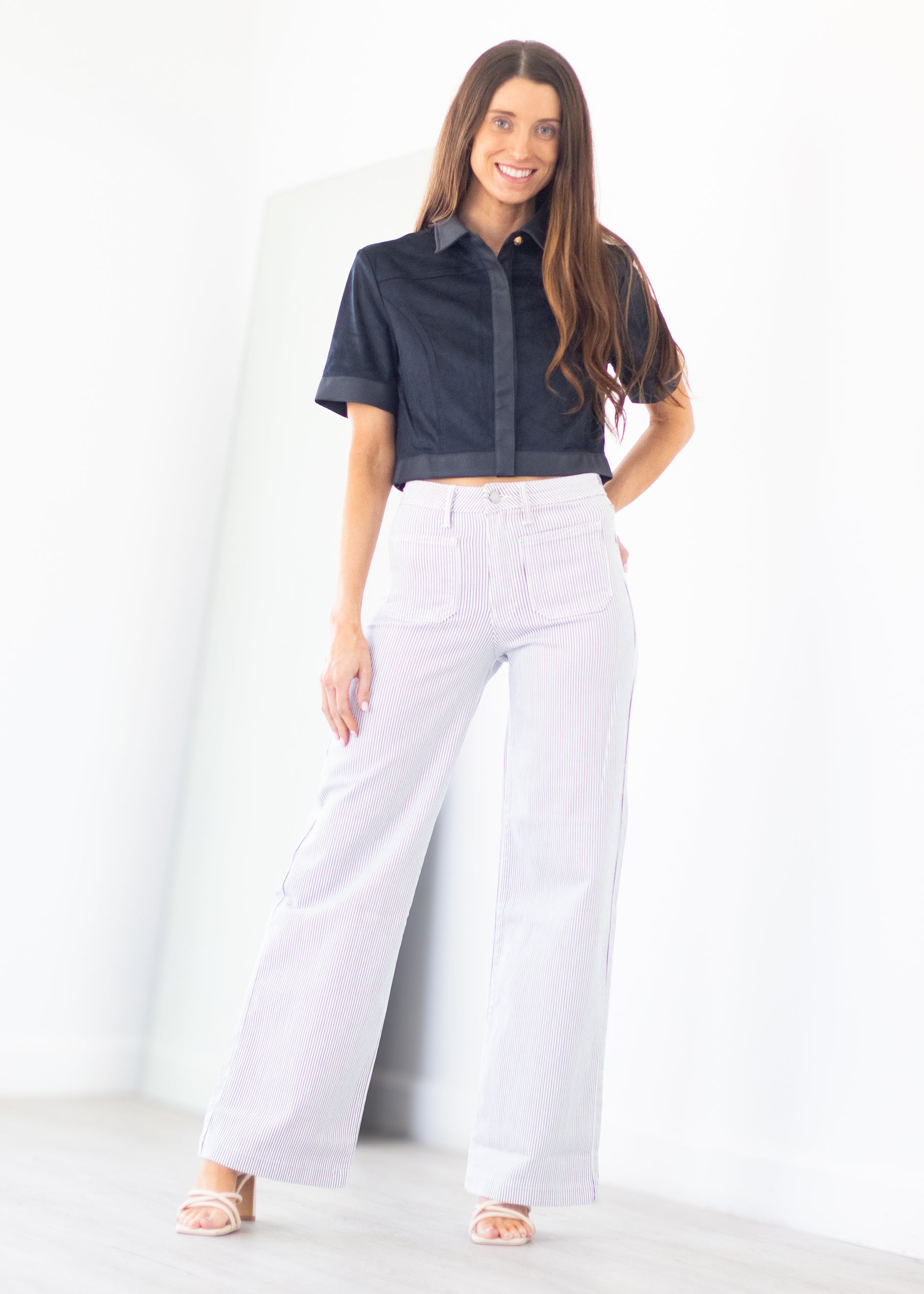A woman modeling a dark navy short-sleeve button-up shirt and white wide-leg pants, standing in a bright white studio setting.