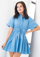 A woman modeling a light blue short-sleeve button-front dress with a gathered waist and flared skirt, photographed indoors against a bright white background.