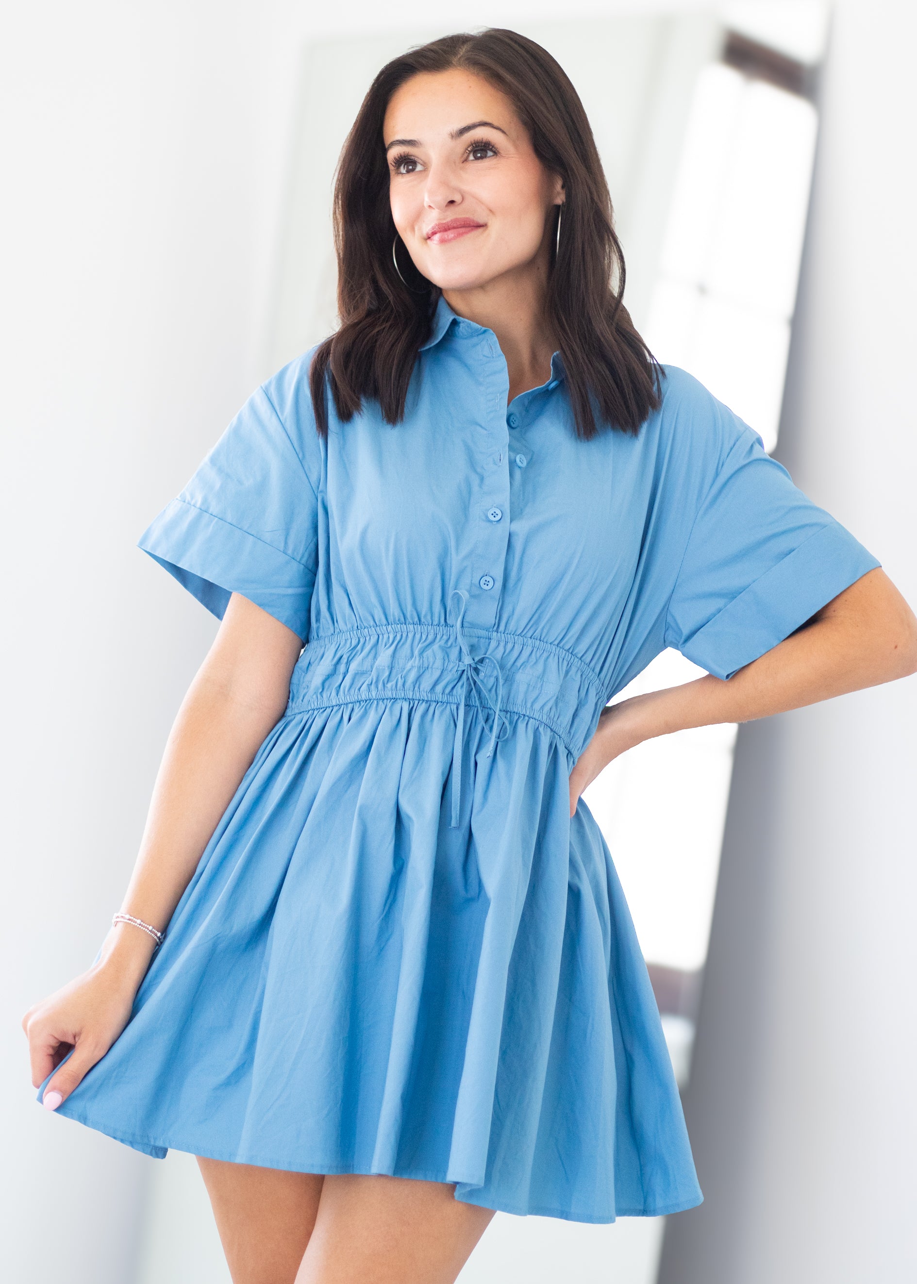 A woman modeling a light blue short-sleeve button-front dress with a gathered waist and flared skirt, photographed indoors against a bright white background.
