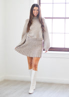 A woman modeling a light beige ribbed knit turtleneck sweater and matching pleated mini skirt, paired with white knee-high boots, standing in a bright room with a window.