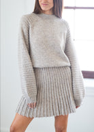 A woman wearing a light beige knit sweater and matching ribbed knit pleated skirt, standing in a bright indoor setting.