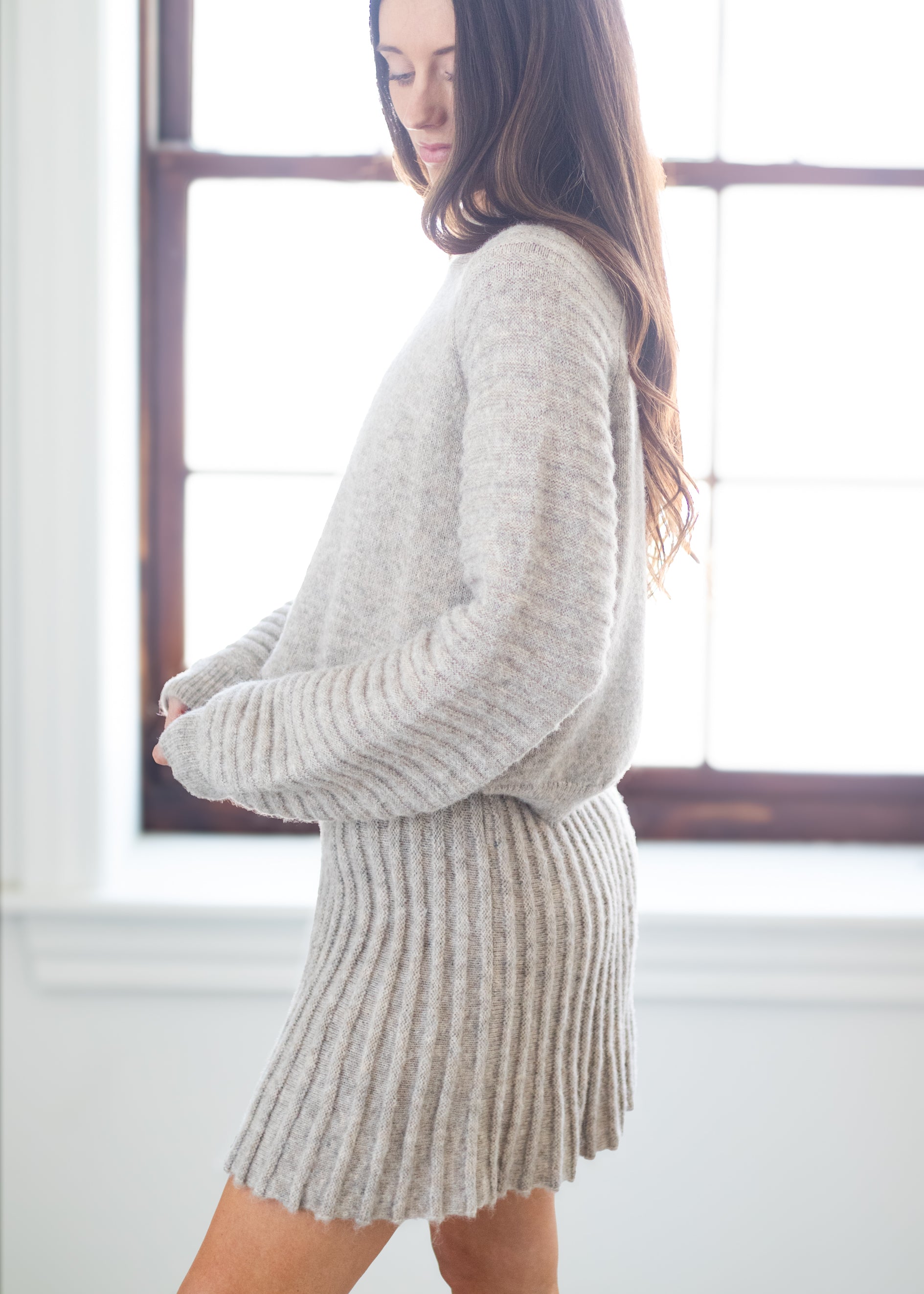 A woman wearing a light heather gray ribbed knit sweater and matching pleated mini skirt, standing in a bright room with a window behind her.