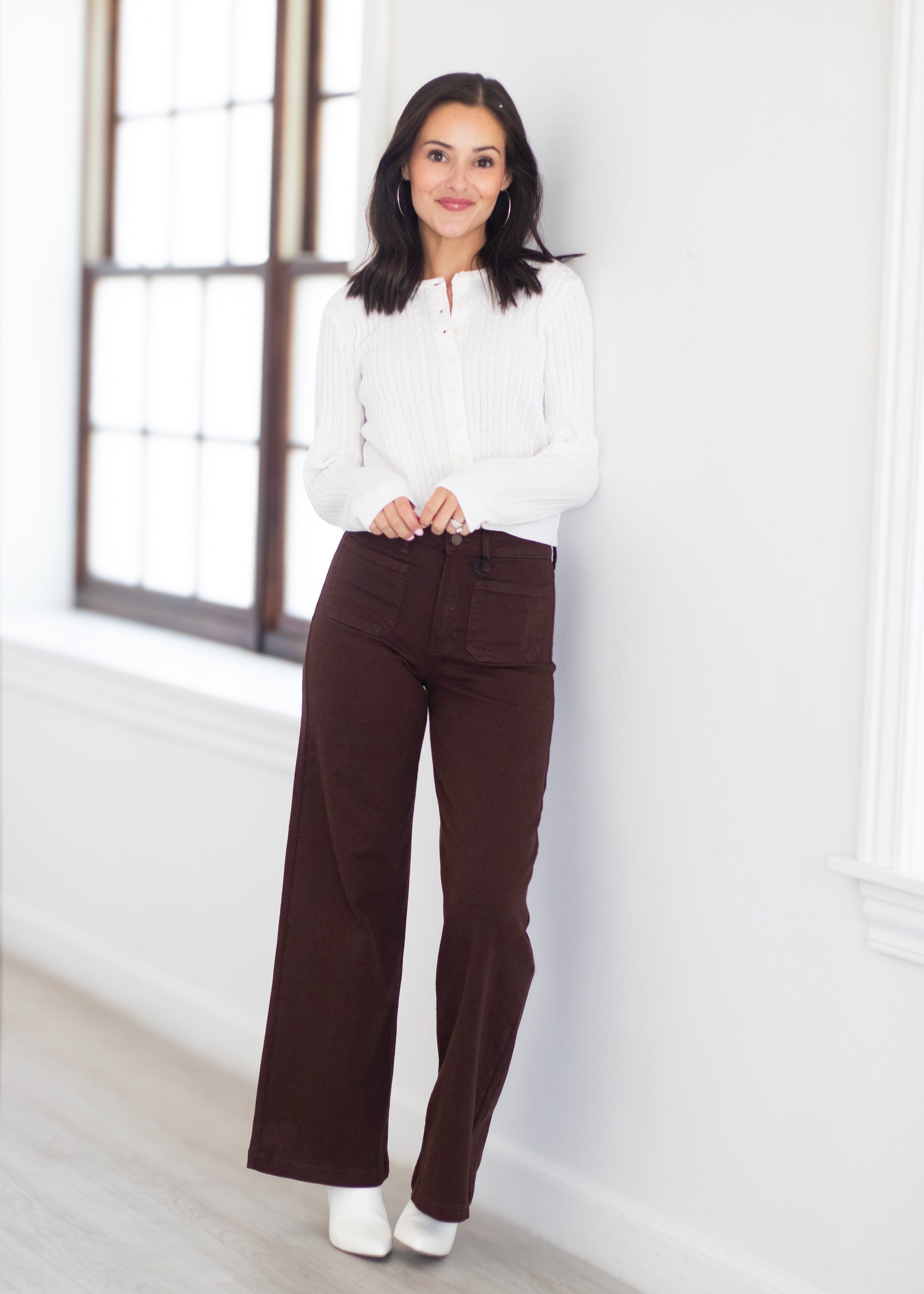 A woman stands indoors wearing a white ribbed long-sleeve top and dark brown wide-leg pants, posed against a white wall near a window.