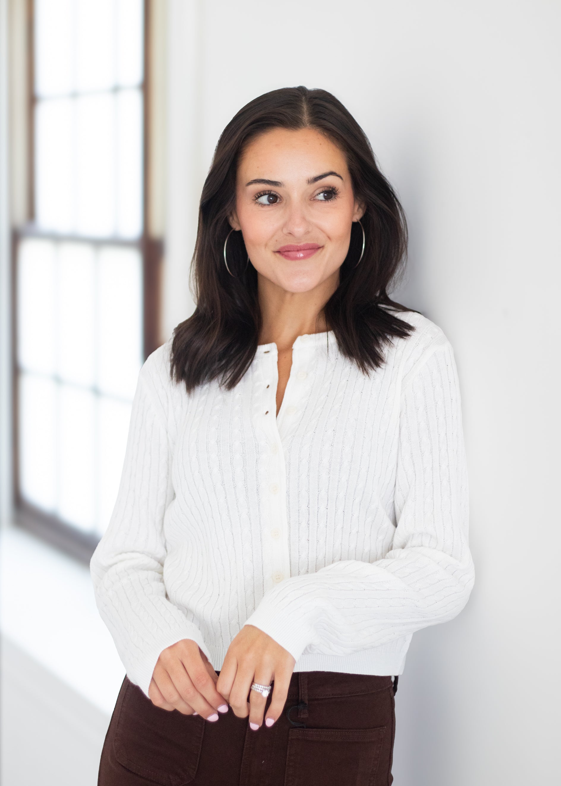 A woman with dark shoulder-length hair wearing a white ribbed long-sleeve button-front top and dark brown pants, standing indoors against a white wall near a window.