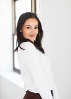 A woman with long dark hair wearing a white ribbed knit sweater and brown pants, looking back over her shoulder in a brightly lit room with a window.