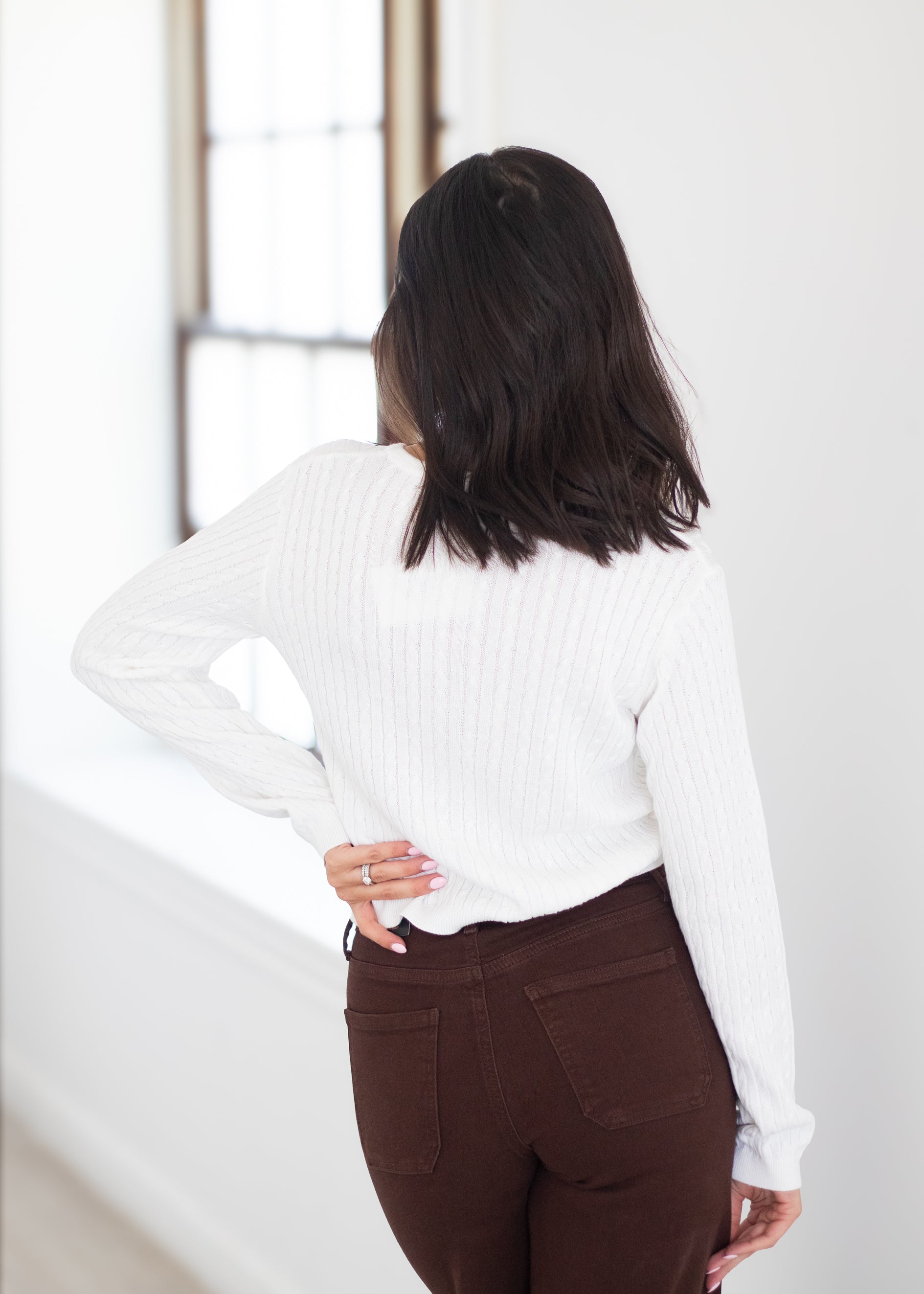 A woman shown from behind wearing a white ribbed knit long-sleeve sweater and dark brown pants in a bright, minimalist room.