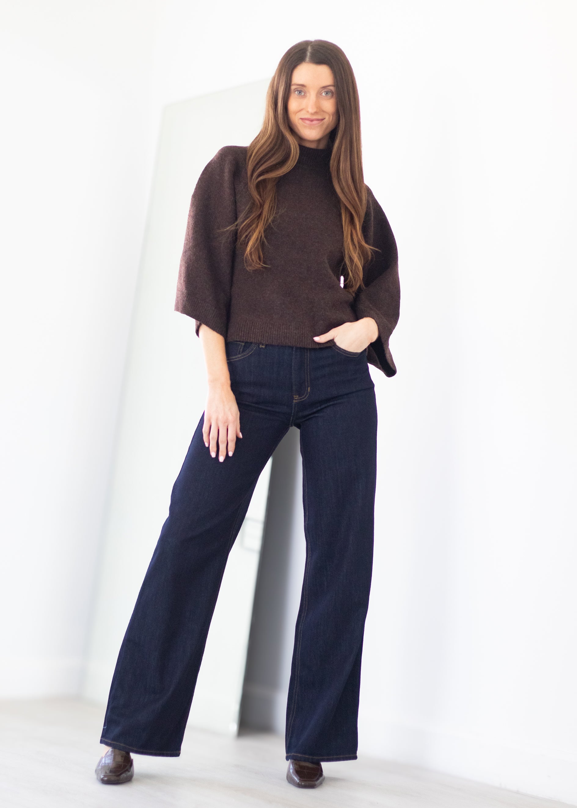 A woman modeling a dark brown fuzzy turtleneck poncho sweater and dark blue wide-leg jeans, standing in a bright white room.