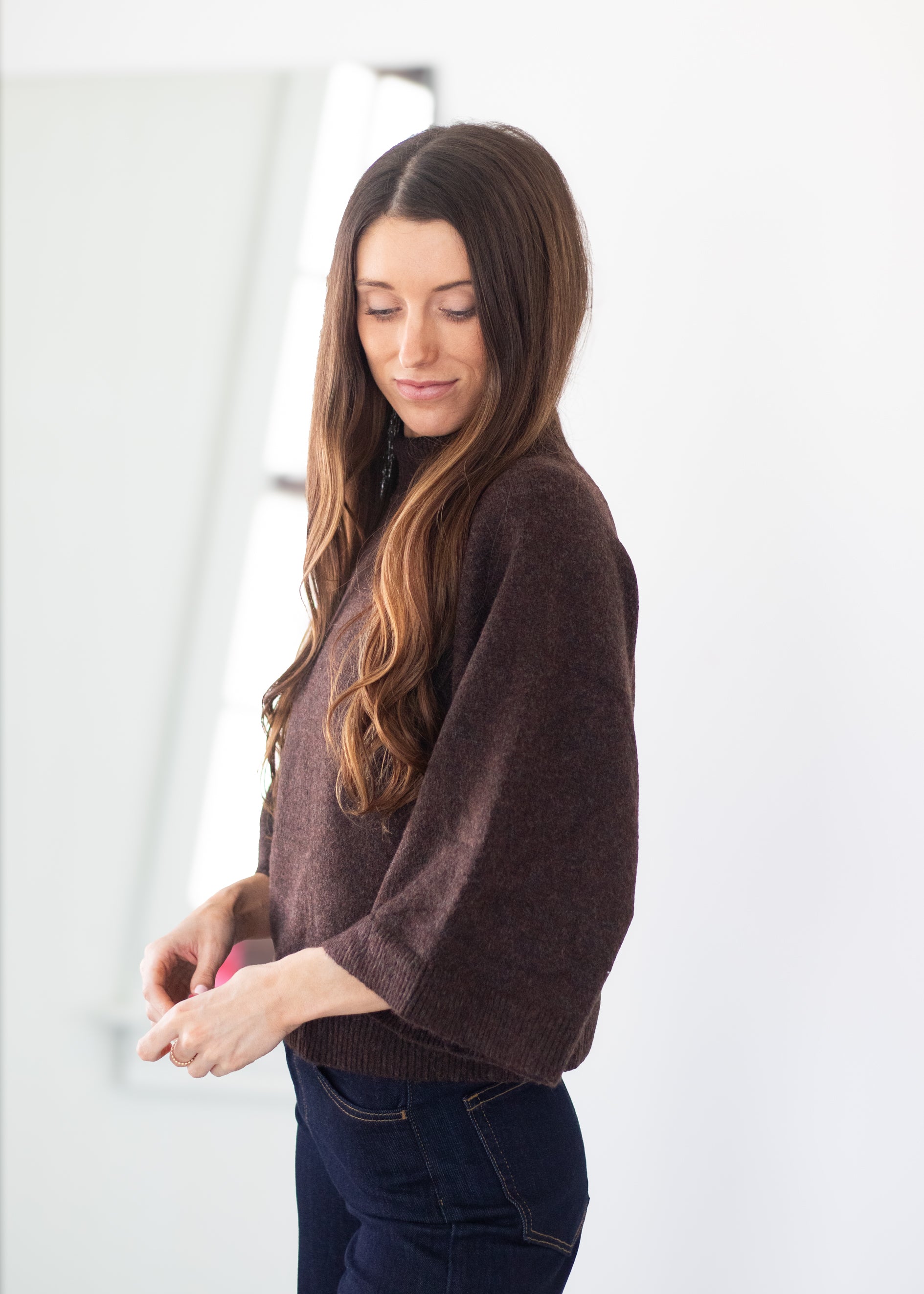 A woman with long brown hair is wearing a dark brown knit sweater and dark blue jeans, standing in a brightly lit white room.