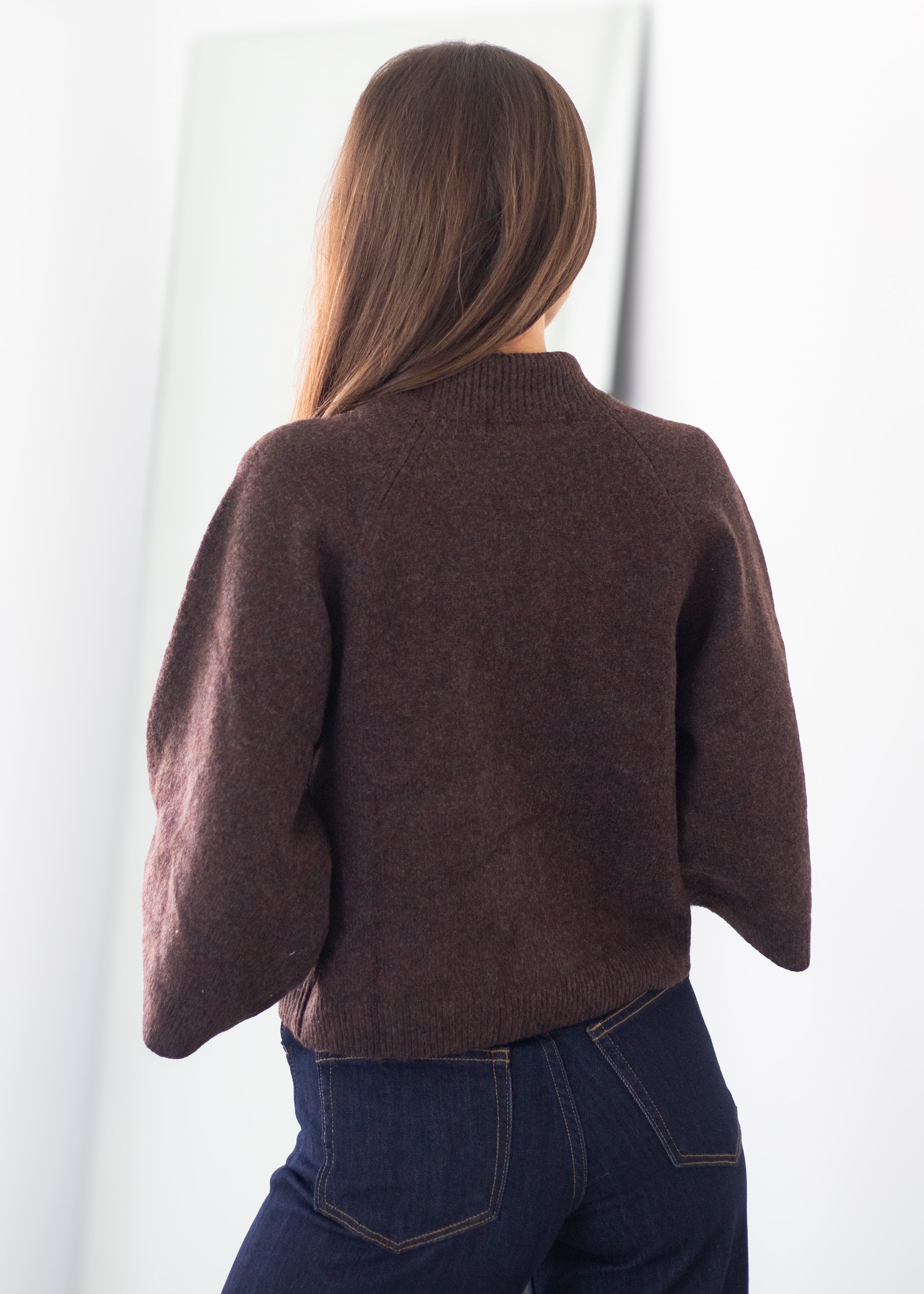 A woman viewed from behind wearing a dark brown cropped knit sweater with wide draped sleeves and dark blue jeans, standing against a bright white background.