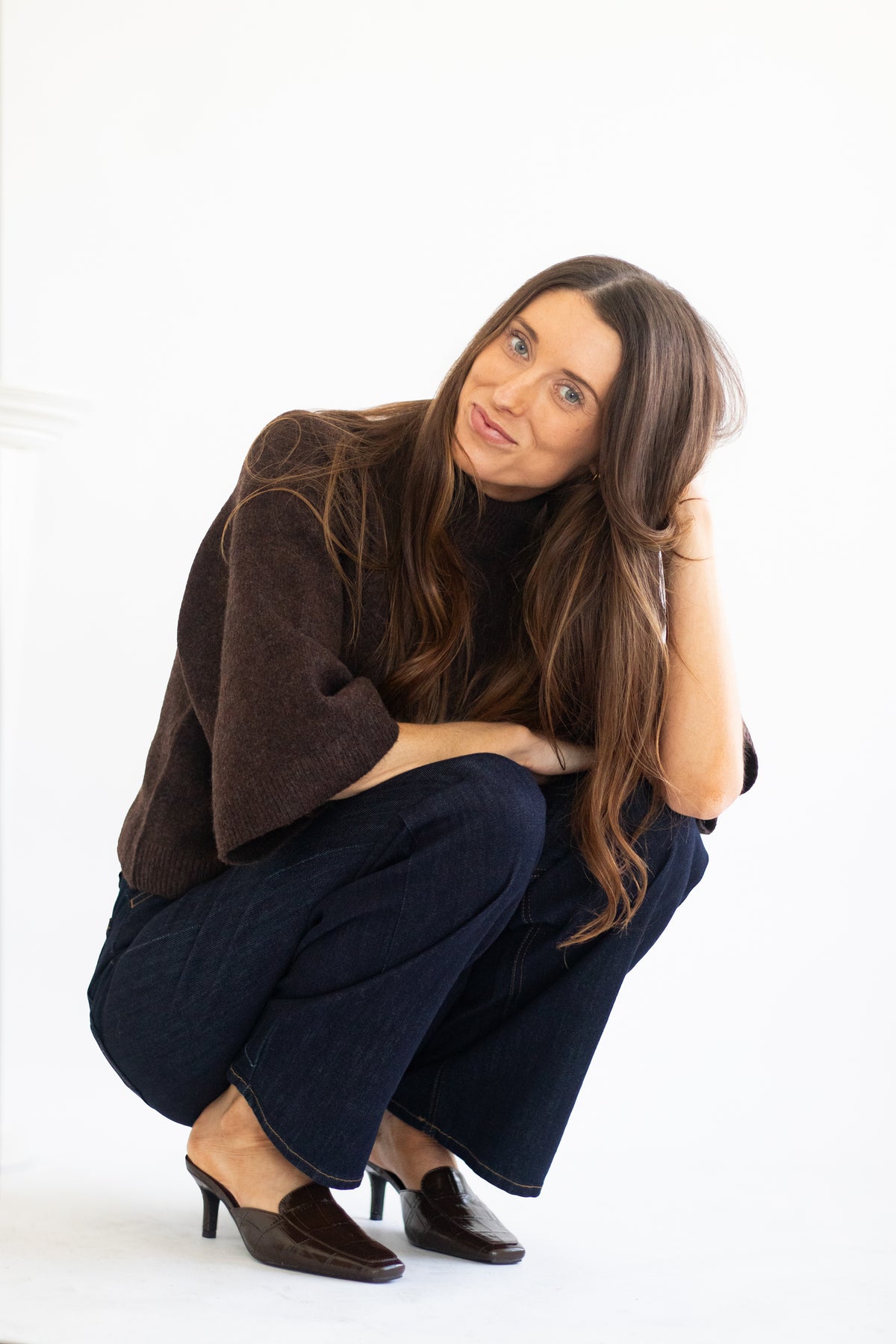 A woman with long brown hair crouches in a studio setting, wearing a dark brown knit cardigan, dark blue wide-leg jeans, and dark brown pointed-toe mule heels against a plain white background.