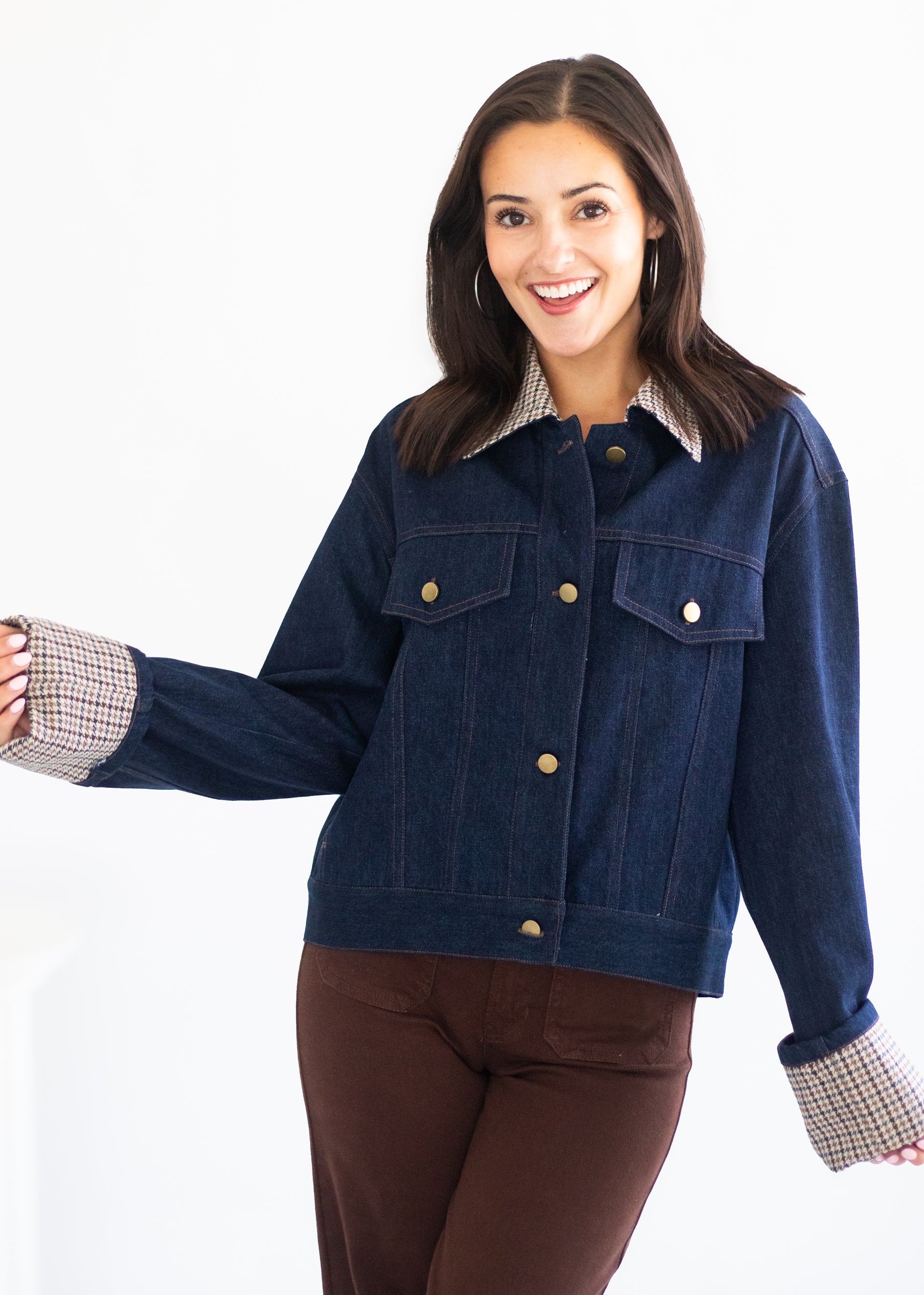 A woman modeling a dark blue denim jacket with a contrasting beige and white checkered collar and cuffs, paired with brown pants, against a plain white background.