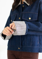 A close-up of a person wearing a dark blue denim jacket with a contrasting black, white, and brown houndstooth patterned lining on the collar and cuffs, paired with brown pants against a white background.