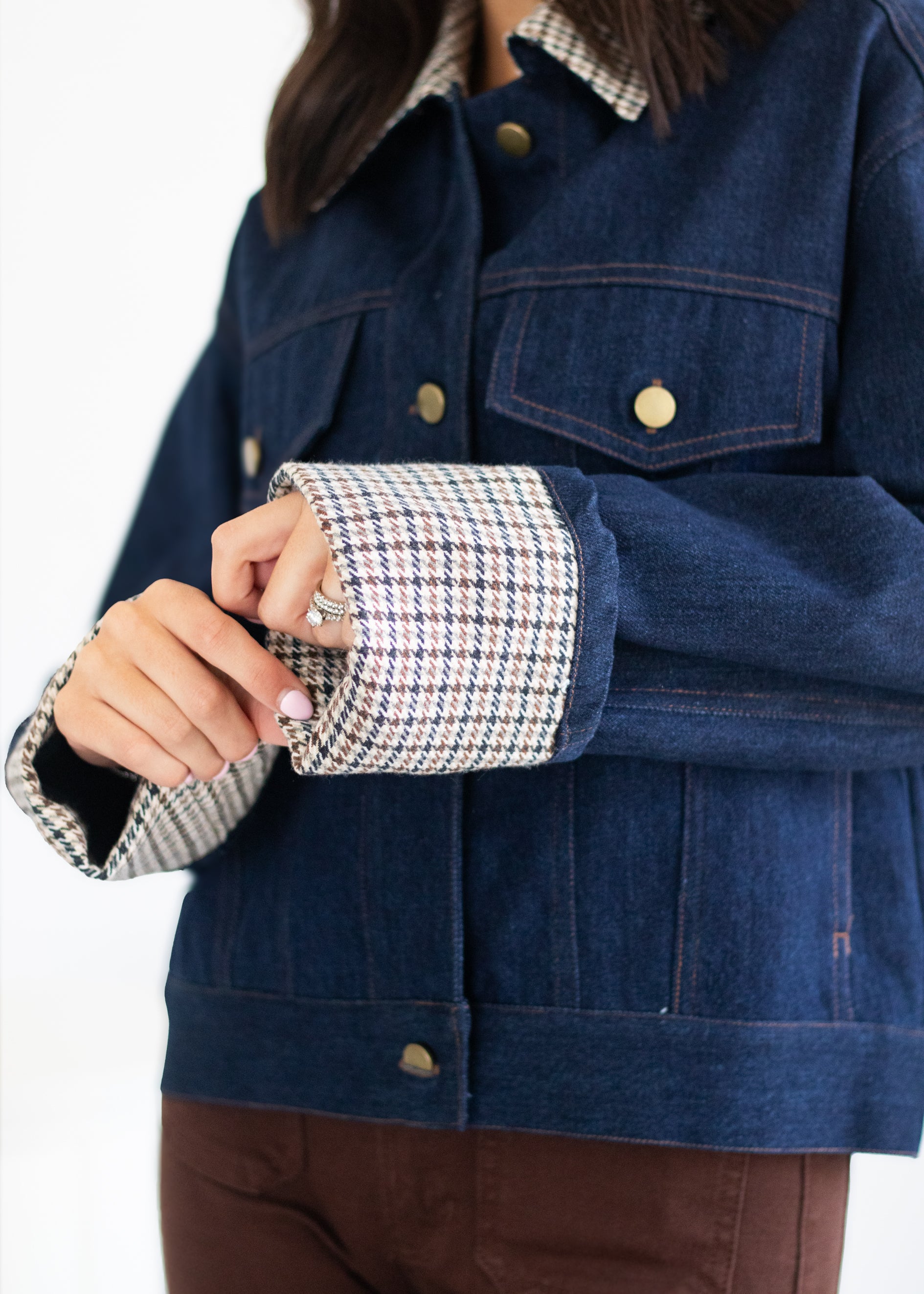 A close-up of a person wearing a dark blue denim jacket with a contrasting black, white, and brown houndstooth patterned lining on the collar and cuffs, paired with brown pants against a white background.