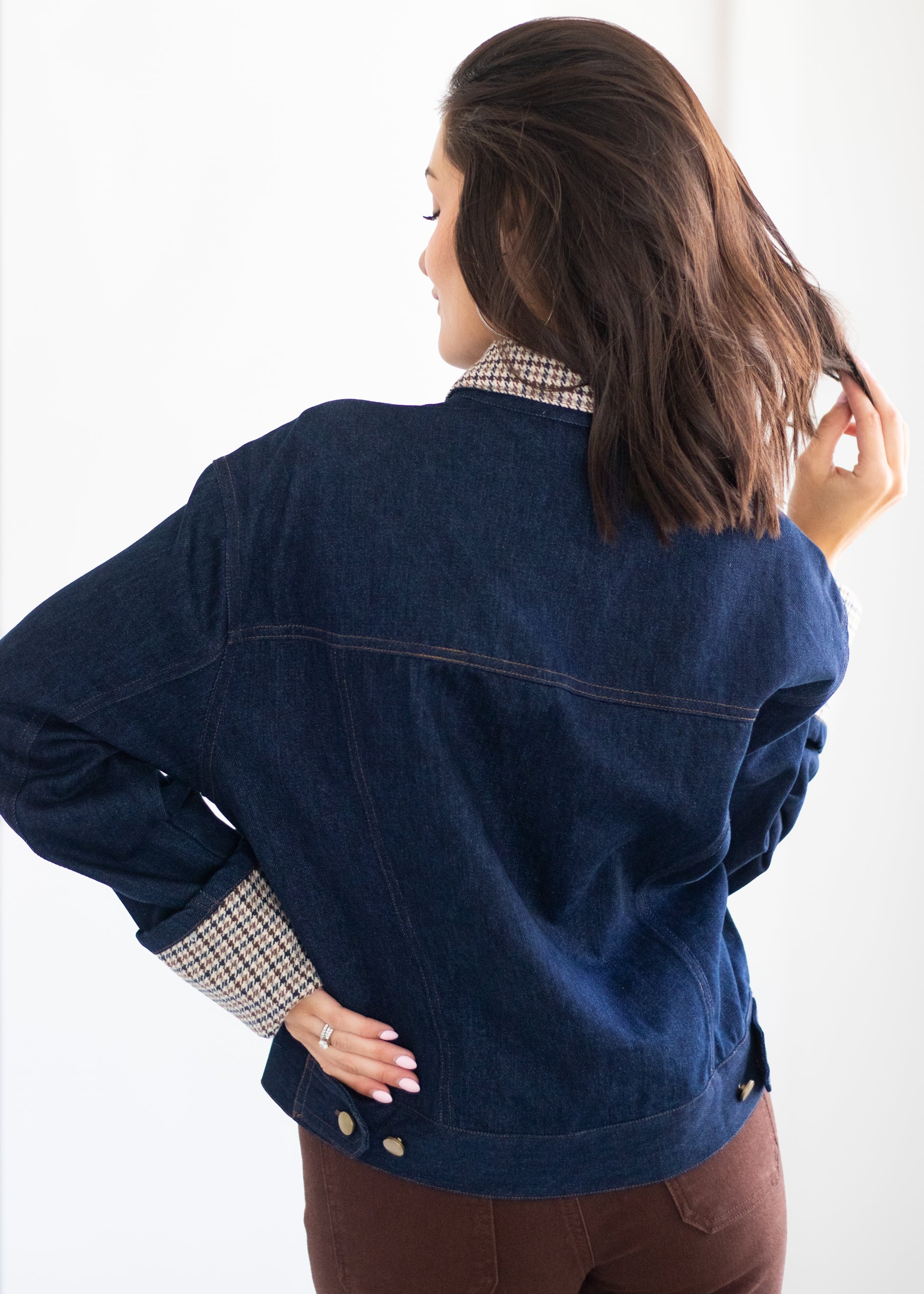 A woman shown from the back wearing a dark blue denim jacket over a beige and white checkered shirt, paired with brown pants, against a plain white background.