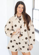 A woman modeling a matching beige two-piece set with large dark brown polka dots, featuring a cropped button-front jacket and coordinating shorts, set against a bright, minimalist indoor background.
