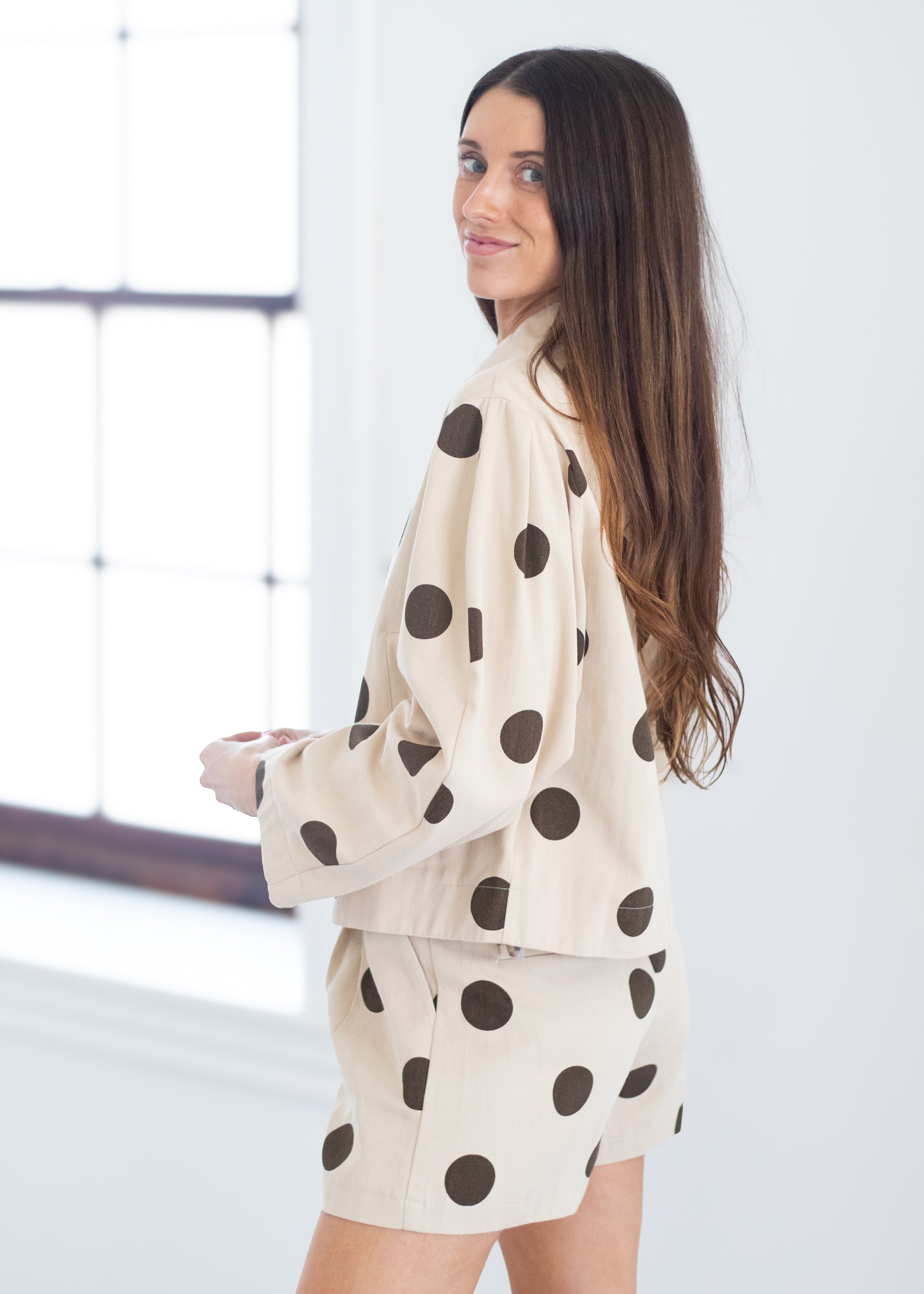 A woman with long brown hair is shown from the back-three-quarter view wearing a matching cream-colored pajama set with dark brown polka dots. The set includes a long-sleeve top and shorts, photographed in a bright, minimalist room with a large window.