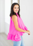A woman wearing a bright pink sleeveless ruffled top and light blue jeans, standing in a bright white indoor setting.