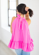A woman shown from behind wearing a bright pink halter top with a ruffled hem, paired with light blue jeans in a bright indoor setting.