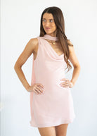 A woman modeling a sleeveless light pink dress with a draped neckline and a keyhole cutout at the collar, set against a plain white studio background.