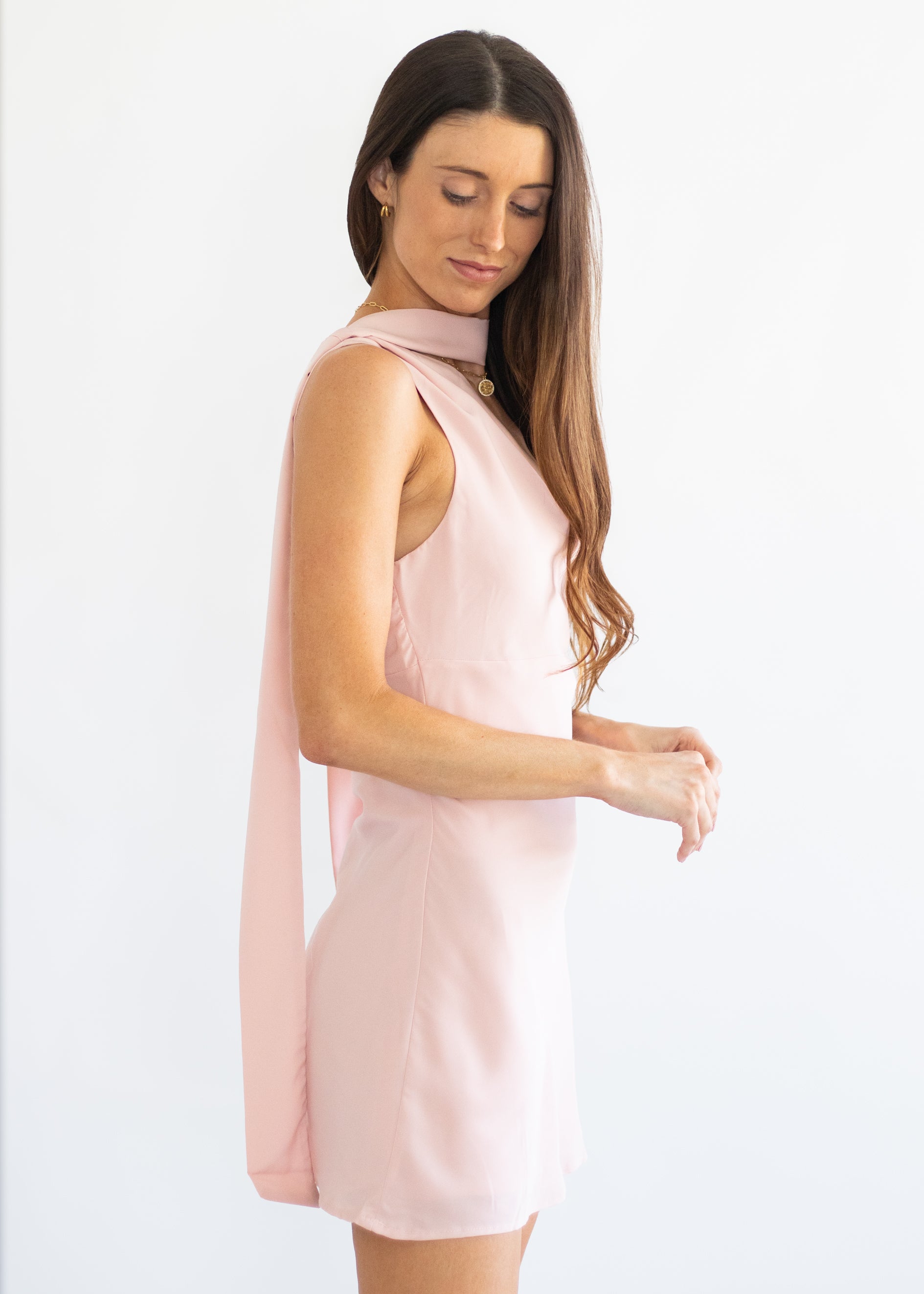A woman modeling a sleeveless light pink dress with a draped high collar and a long flowing cape-like back drape, photographed against a clean white studio background.