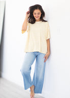 A woman wearing a yellow and white striped short-sleeve t-shirt and light blue wide-leg jeans, standing against a white wall.